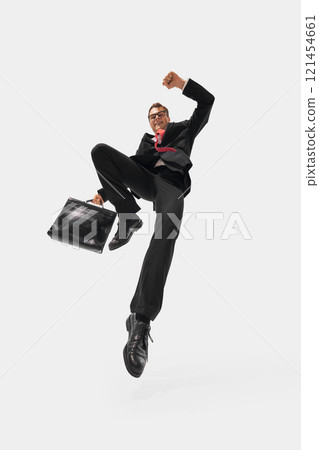 Business professional in black suit and red tie jumping energetically, raising fist and holding briefcase against white studio background. 121454661