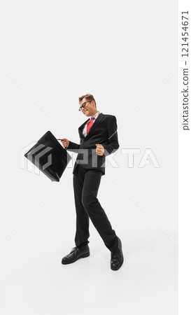Man in sharp black suit and red tie striking exaggerated confident pose while holding briefcase in hand against white studio background. 121454671