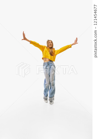 Woman in vibrant yellow sweater and casual blue jeans standing confidently with arms stretched wide and relaxed posture against white studio background. 121454677