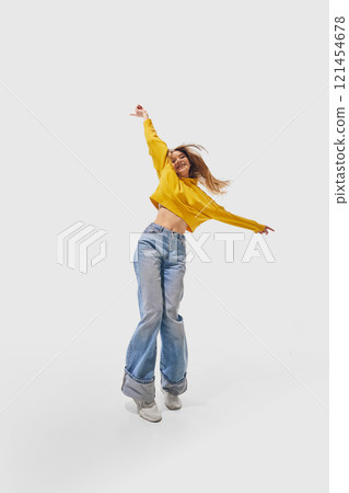 Student in yellow sweater and blue jeans extending arms joyfully while leaping mid-air with hair cascading around her against white studio background. Student in yellow sweater and blue jeans extending arms joyfully while leaping mid-air with hair cascading around her against white studio background. 121454678