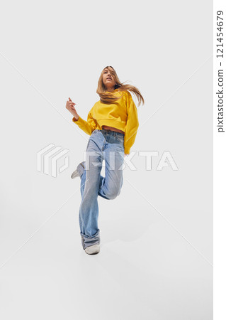 Woman wearing yellow sweater and blue jeans energetically jumping with loose hair flowing freely against white studio background. 121454679