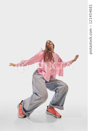 Woman squats slightly, while dancing dressed in casual pink and denim, with expressive confidence against white studio background. 121454681