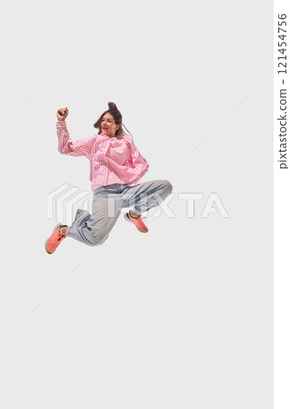 Woman in casual pink and denim leaps mid-air with joyful expression, showcasing energy and motion against white studio background. 121454756
