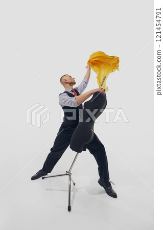 Man, stylist, fashion designer pulls yellow fabric draped over black mannequin with purposeful motion against white studio background. 121454791