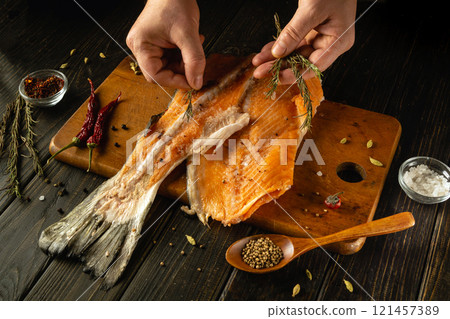 Hands skillfully separate herbs from a beautifully smoked salmon fillet on a rustic wooden board. Spices and salt surround the fish, enhancing the culinary art of preparation 121457389