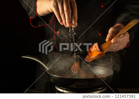 Chef expertly adds a pinch of salt to sizzling pieces of squash in a frying pan. The warm kitchen is filled with aromatic steam and promise of delicious food, demonstrating culinary skill 121457391