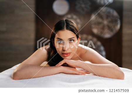 Close-up portrait of young woman lying on table with looking at camera preparing for relax massage procedure in wellness center. Happy beautiful brunette female awaiting beauty treatment at spa salon. 121457509