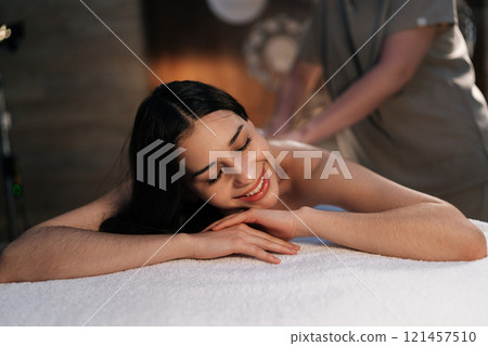 Portrait of smiling young woman relaxing during back massage in spa with soft light. Beautiful naked female with perfect skin getting relaxing massage. Concept of healthy lifestyle and body care 121457510