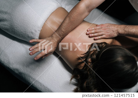High-angle view of masseur giving back massage to female client lying on massage table in modern spa center with dark interior. Beautiful young woman enjoying relaxing treatment in spa room. High-angle view of masseur giving back massage to female client lying on massage table in modern spa center with dark interior. Beautiful young woman enjoying relaxing treatment in spa room. 121457515