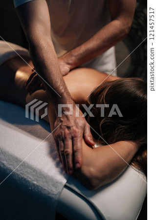 Vertical cropped shot of massage therapist performing deep tissue technique on female shoulder, targeting muscle tension and promoting wellness during bodywork session in spa salon with dark interior 121457517