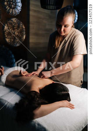 Vertical shot of focused masseuse giving relaxing back massage to relaxed young woman lying on massage table in spa, creating tranquil and rejuvenating experience. Concept of professional massage. Vertical shot of focused masseuse giving relaxing back massage to relaxed young woman lying on massage table in spa, creating tranquil and rejuvenating experience. Concept of professional massage. 121457519