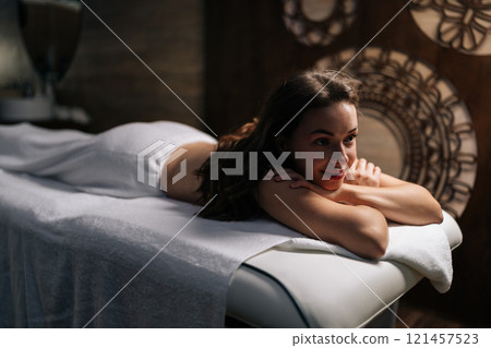 Young woman lying comfortably on massage table, wrapped in soft white towel, enjoying moment of tranquility while waiting for massage in luxurious spa center. Concept of rest with spa treatment. 121457523