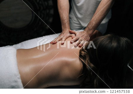 Top view of masseur performing relaxing back massage on female client lying on massage table, focusing on shoulder blades and applying oil in spa center with dark interior. Concept of luxury massage. 121457538