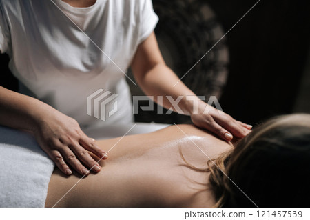 Cropped shot of masseuse giving relaxing back massage to young unrecognizable woman lying on massage table in spa with dark interior, creating tranquil and soothing atmosphere, close-up. 121457539