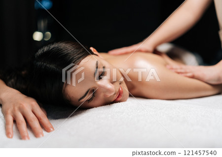 Close-up portrait of beautiful calm young woman client relaxing during back massage in spa salon with dark interior. Professional massage therapist providing therapeutic back massage to relaxed female 121457540