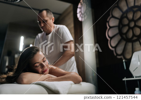 Beautiful young woman relaxing with hand massage at beauty spa lying with closed eyes. Professional masseur giving back massage to female client experiencing therapeutic wellness in beauty parlor. Beautiful young woman relaxing with hand massage at beauty spa lying with closed eyes. Professional masseur giving back massage to female client experiencing therapeutic wellness in beauty parlor. 121457544
