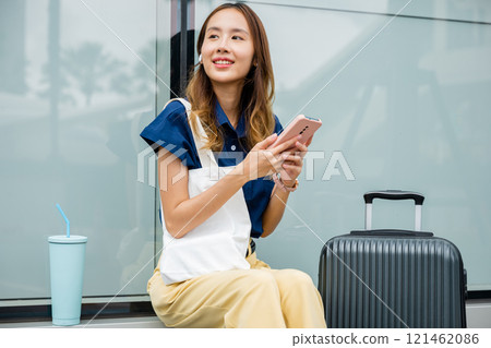 A young woman sitting and relaxing with her suitcase, using her mobile phone to browse social media and stay connected while waiting for her flight at the airport. She is trendy and modern. 121462086