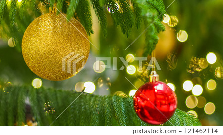 a gold glittery ornament and a shiny red bauble hanging from green branches of a Christmas tree. The background is softly blurred with warm yellow lights, creating a festive and cozy holiday ambiance 121462114