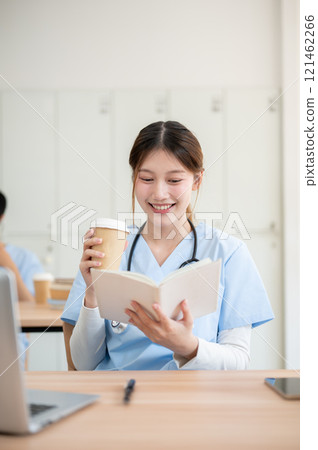 An Asian female medical student in scrubs is sipping a hot drink while reading in a library. An Asian female medical student in scrubs is sipping a hot drink while reading in a library. 121462266