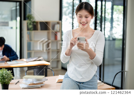 A smiling Asian female college student reading texts on her smartphone while standing in a library. 121462364