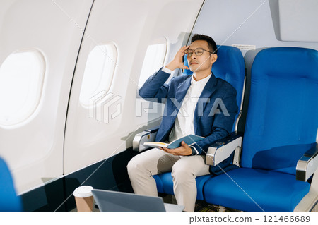 Photo of a frustrated man sitting on an airplane with her head in her hands. Asian Businessman sitting in airplane working laptop and tablet 121466689