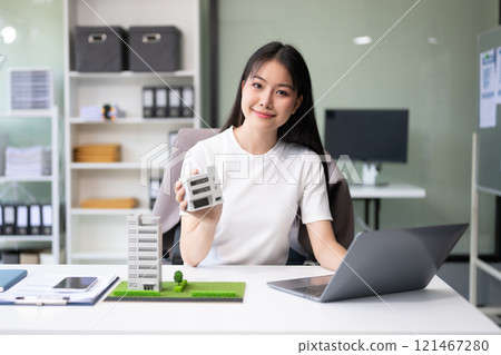 Young real estate agent worker working with laptop and tablet at table in office and small house beside it. 121467280