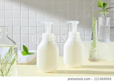 In the center are two white pump bottles no label, surrounded by some glass vases containing a green tea leaves and fern branches, place on a yellow surface, in a bathroom backdrop. Advertising photo. In the center are two white pump bottles no label, surrounded by some glass vases containing a green tea leaves and fern branches, place on a yellow surface, in a bathroom backdrop. Advertising photo. 121468008