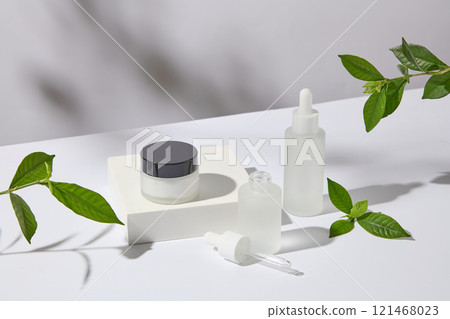 Mockup of a jar and serum bottles no label, set on a white pedestal with green tea leaves - anti-acne and moisturizing benefits. Side view. A photo to promote skincare products Mockup of a jar and serum bottles no label, set on a white pedestal with green tea leaves - anti-acne and moisturizing benefits. Side view. A photo to promote skincare products 121468023