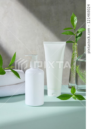 A close-up of a set of unlabeled products, a white tube and a white pump bottle, placed on a blue surface, next to glass vases containing green tea leaves and tea tree branches, in a grey backdrop. A close-up of a set of unlabeled products, a white tube and a white pump bottle, placed on a blue surface, next to glass vases containing green tea leaves and tea tree branches, in a grey backdrop. 121468024