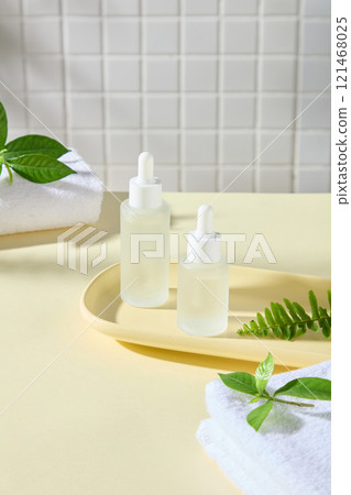 Mockup of dropper bottles placed on a wooden tray, next to some towels, green tea leaves, and fern fronds, in a bathroom setting. Advertising photos for serum products. Mockup of dropper bottles placed on a wooden tray, next to some towels, green tea leaves, and fern fronds, in a bathroom setting. Advertising photos for serum products. 121468025