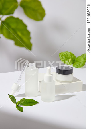 Side angle, shows serum bottles with a cream jar, placed on a white podium, next to some green tea leaves - helps prevent acne and moisturize. Advertisement photo. Side angle, shows serum bottles with a cream jar, placed on a white podium, next to some green tea leaves - helps prevent acne and moisturize. Advertisement photo. 121468026