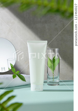 Front view photo for skincare advertising, unlabeled white tube display on a blue surface, next to a glass, green tea leaves, fern branches, a towels, and a mirror, in a grey background. Front view photo for skincare advertising, unlabeled white tube display on a blue surface, next to a glass, green tea leaves, fern branches, a towels, and a mirror, in a grey background. 121468027