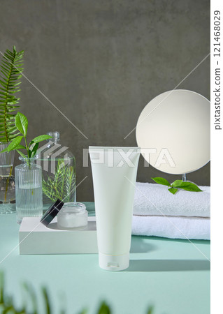 An unbranded white tube standing on a blue surface, next to a cream jar on a podium, glass vases containing green tea leaves, tea trees, and fern branches, in a gray backdrop. Template ad photo. 121468029