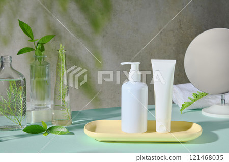 Frontal view of an unbranded white pump bottle and tube placed on a wooden tray, on a blue surface, surrounded by some glass vases containing green tea and tea tree branches in a spa room setting. 121468035