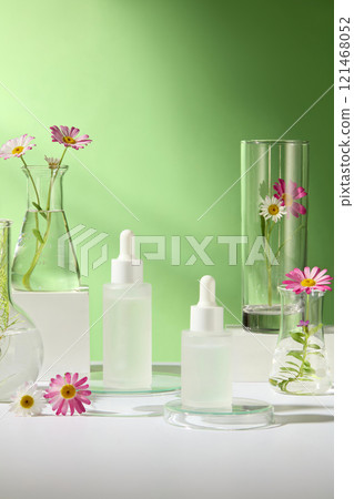 Close-up of an unlabeled mockup of dropper bottles on white and glass podiums, surrounded by some lab glassware with chamomile flower branches, a green backdrop for a cosmetics advertisement photo. 121468052