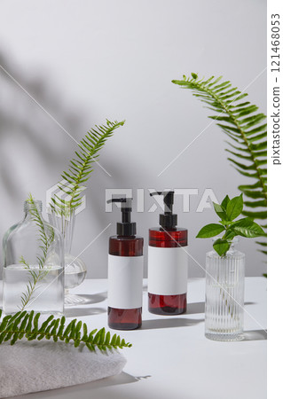 In a white minimalist backdrop, a front view of two dark brown pump bottles is placed on a white surface, next to towels and some glass vases containing ferns and green tea branches. Advertising photo 121468053