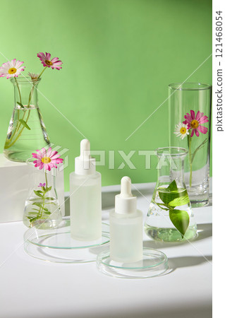 A green backdrop for a skincare ad photo, high view of an unlabeled mockup of dropper bottles on glass podiums, next to lab glassware with chamomile branches and green tea leaves on a white platform. A green backdrop for a skincare ad photo, high view of an unlabeled mockup of dropper bottles on glass podiums, next to lab glassware with chamomile branches and green tea leaves on a white platform. 121468054