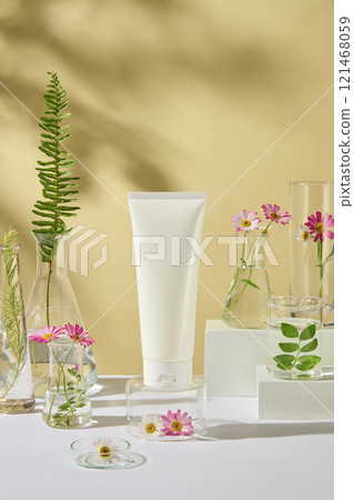 Close-up view of an unbranded cosmetics tube mockup, displayed on a glass podium, decorated by fresh tea trees, ferns, and chamomile branches. Mockup for vegan cosmetics advertisement photo. 121468059