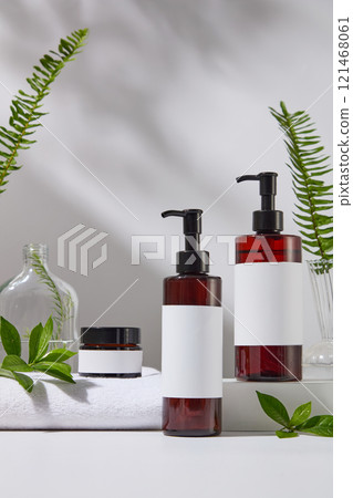 A mockup of a jar and two pump bottles, shown in front of a white platform, surrounded by towels and glass vases holding ferns, tea plants, and green tea branches. Skincare products advertising photo. 121468061