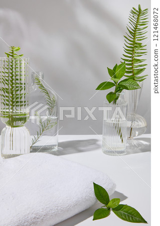 Spa concept with a few glass vases filled with some fresh green teas, ferns, and tea tree leaves, next to empty towels for display products, against a light background. 121468072