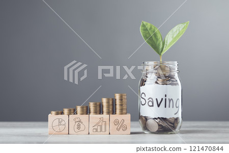 A glass jar filled with coins and a sprout labeled Saving, wooden blocks marked 2025, and neatly stacked coins on a table, symbolizing financial growth and future aspirations against a gray backdrop A glass jar filled with coins and a sprout labeled Saving, wooden blocks marked 2025, and neatly stacked coins on a table, symbolizing financial growth and future aspirations against a gray backdrop 121470844