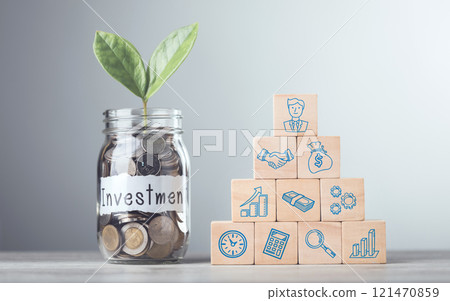 A Glass jar with coins and a sprout labeled 'Investment,' alongside stacked wooden blocks with financial symbols, placed on a table against a gray background, representing savings growth 121470859