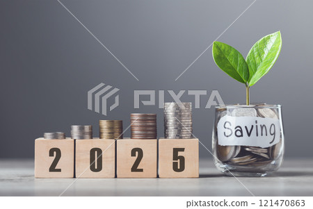 A glass jar filled with coins and a sprout labeled Saving, wooden blocks marked 2025, and neatly stacked coins on a table, symbolizing financial growth and future aspirations against a gray backdrop 121470863