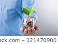 Glass jar filled with coins, with a small seedling growing in the center, placed on a table against a dark grey backdrop. The image represents the concept of saving and financial growth for future 121470900