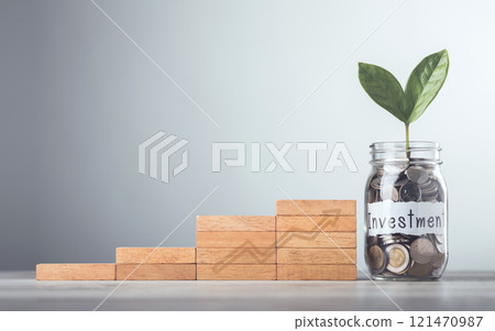 Glass jar filled with coins and a sprout labeled "Investment," placed beside stacked wooden blocks arranged like stairs with interest symbols, symbolizes financial growth, incremental savings 121470987