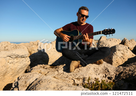 A man in a dark t-shirt playing acoustic guitar on a rocky area by the sea A man in a dark t-shirt playing acoustic guitar on a rocky area by the sea 121472511