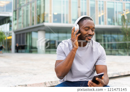 Music song concept. Good start day. Happy African American man listening modern hit on street in city. Person guy wearing headphones enjoy listening favorite music on phone. People lifestyle joy 121475145