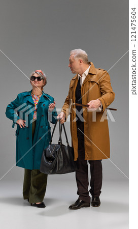Woman wearing teal trench coat, holding large black bag, and man in mustard coat with cane, both sharing lighthearted moment against gray studio background. Woman wearing teal trench coat, holding large black bag, and man in mustard coat with cane, both sharing lighthearted moment against gray studio background. 121475604