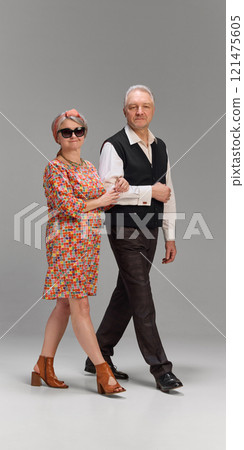 Senior couple walking arm in arm, woman in bright dress and open-toed shoes, man in dark plaid trousers and black vest against gray background. 121475605