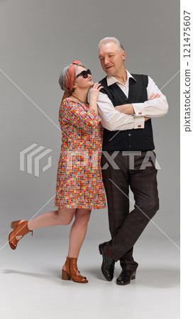 Woman in patterned dress leaning slightly with playful expression, man standing confidently with arms crossed against grey background. 121475607